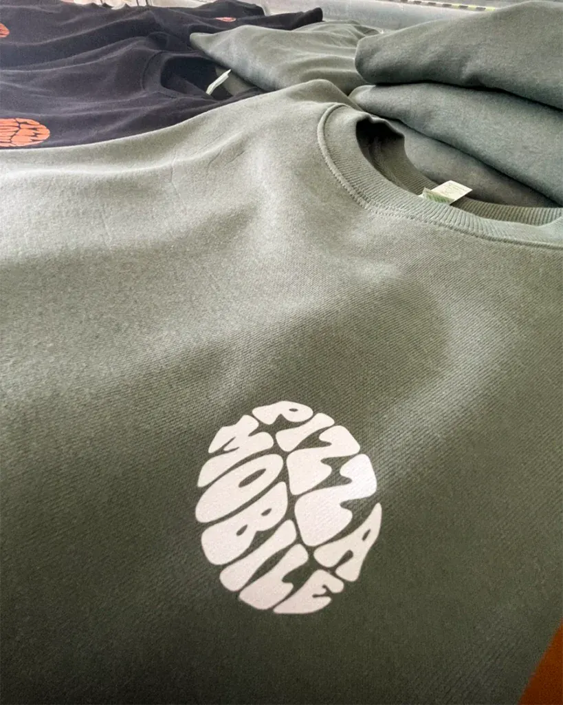 Camisola verde com logótipo "Pizza Mobile" em branco, empilhada com outras roupas.