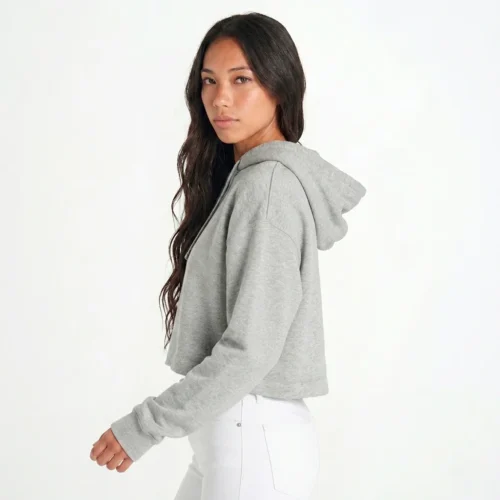 Mulher com cabelo castanho, de costas viradas para a direita e virada para a câmara, vestindo hoodie cropped cinzento e calças brancas.