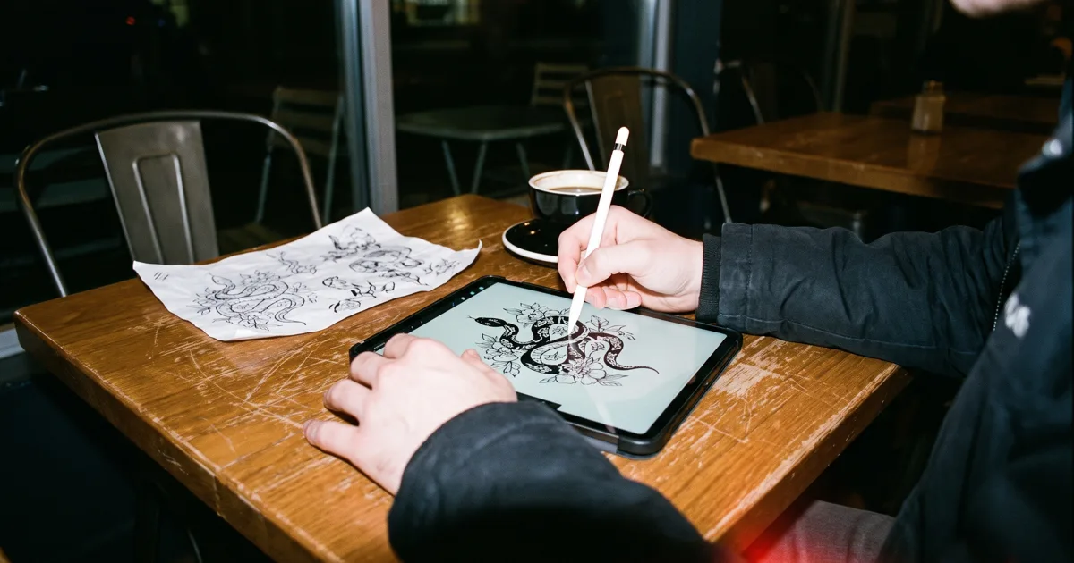 Illustrator's hands drawing a snake with flowers on an iPad, with the sketch on paper and a coffee next to it, on a coffee shop table at night