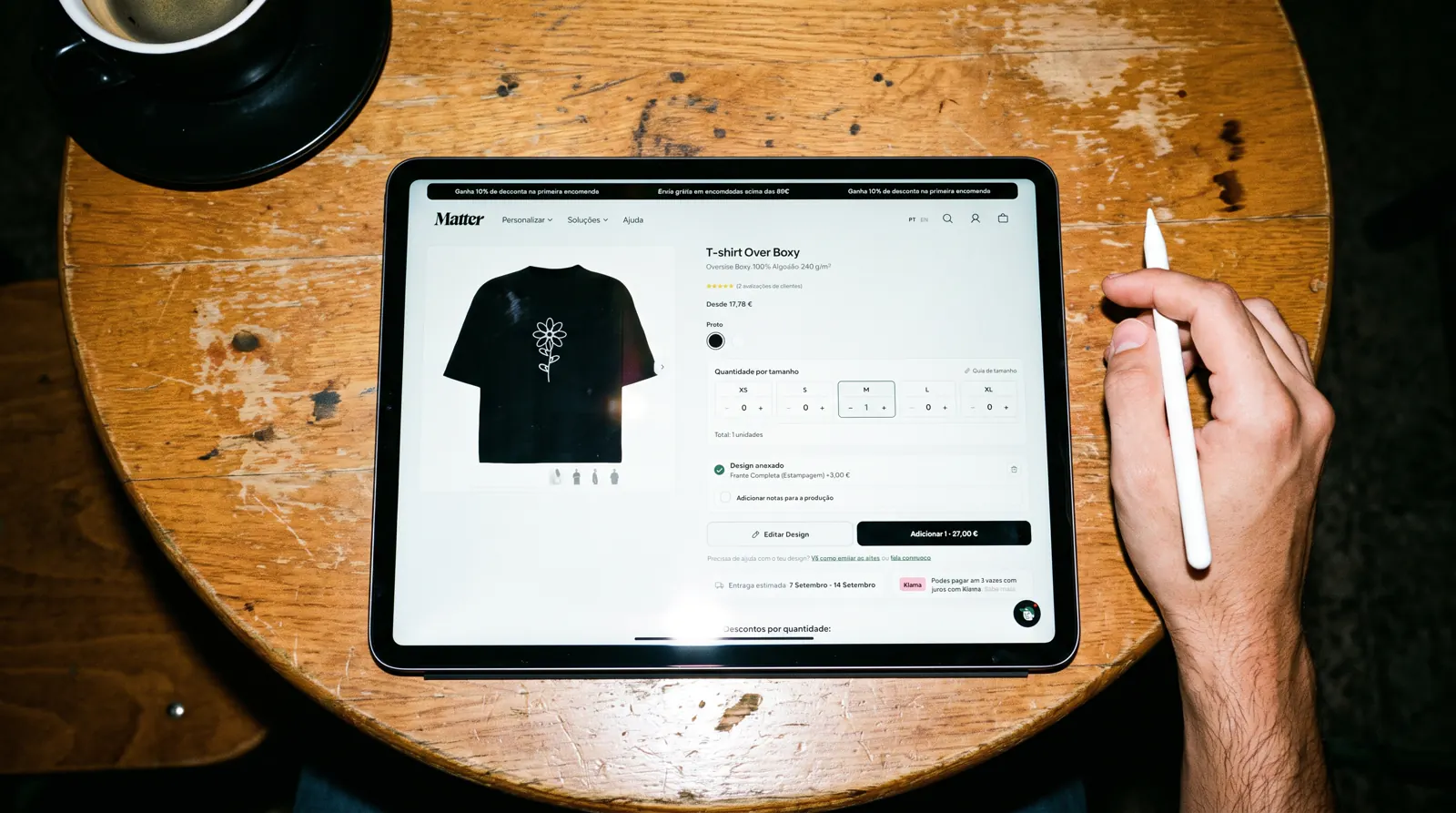 iPad on a coffee shop table showing the Matter Studio customizer with a black oversized t-shirt featuring a white line-art flower, one unit in the cart